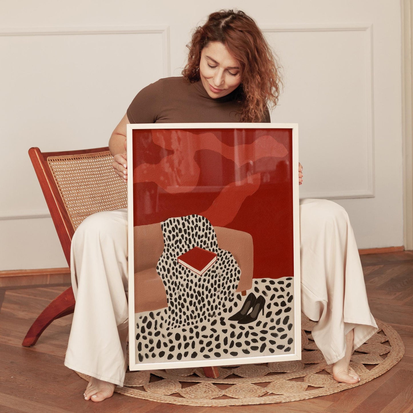 A woman is sitting on a chair and holding a framed print with a red background and a black and white pattern.