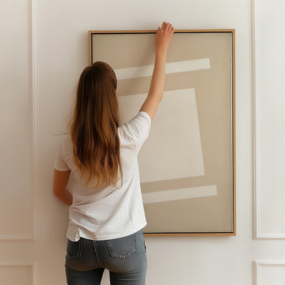 A woman with long brown hair is standing in front of a large picture frame, reaching up to hang it on a white wall.