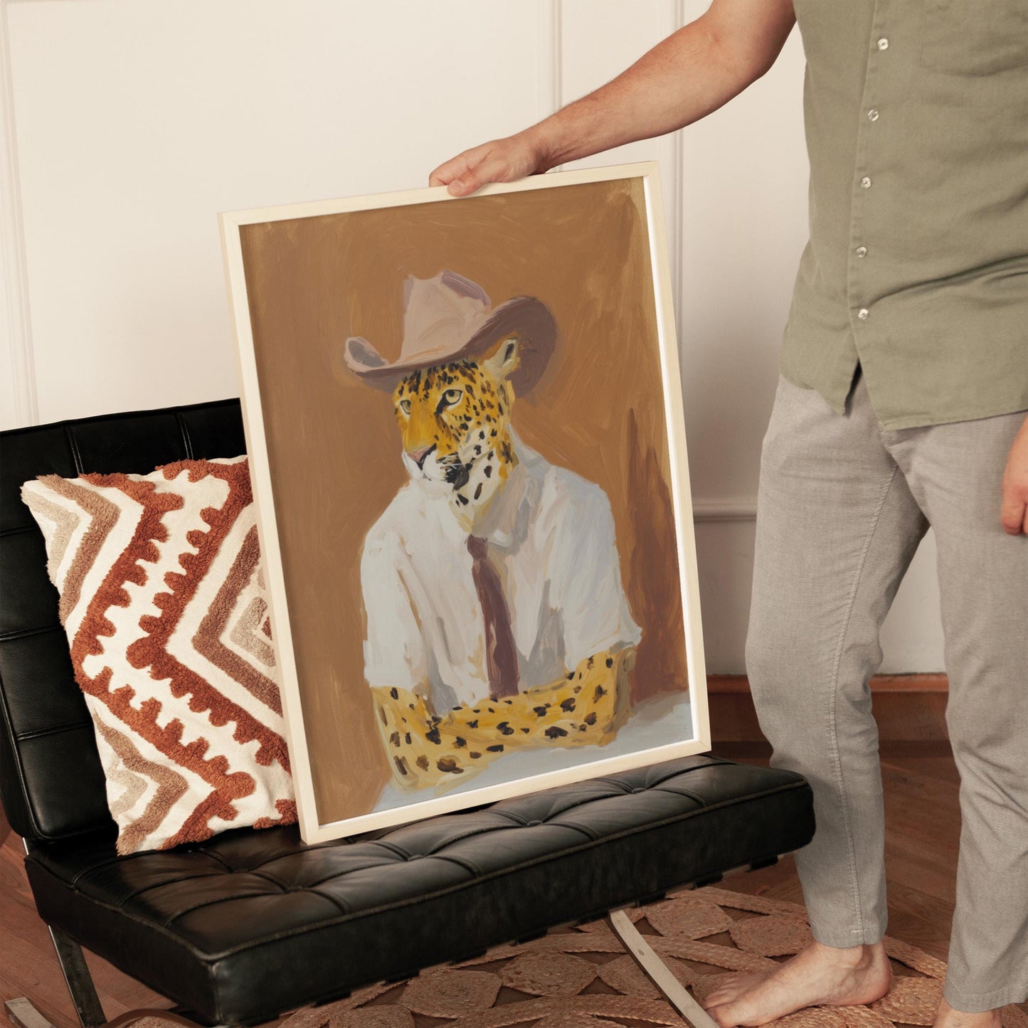 A person is holding up a framed painting of a leopard wearing a white shirt and a cowboy hat.