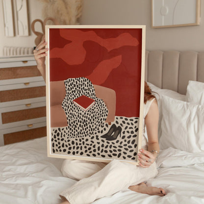 A person is sitting on a bed holding up a framed print with a red background and a black and white leopard print design.