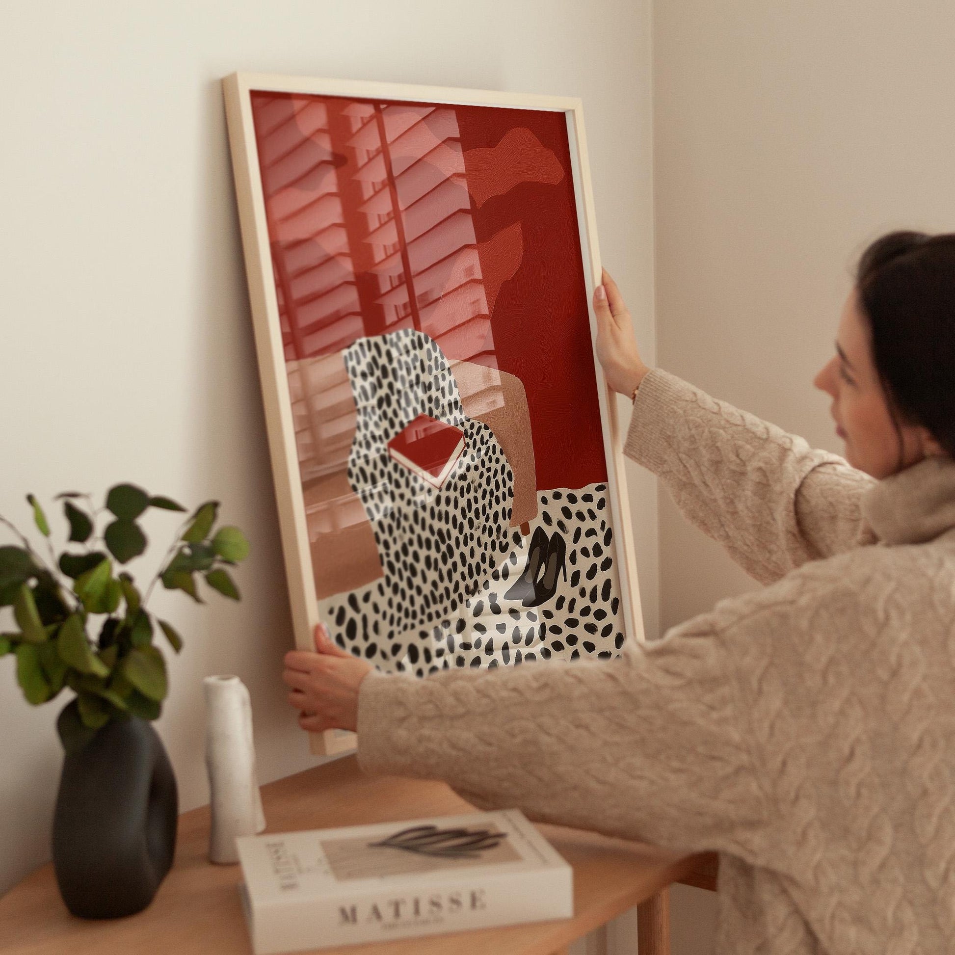 A woman is hanging a framed artwork on a wall, which features a red background with a black and white leopard print design.