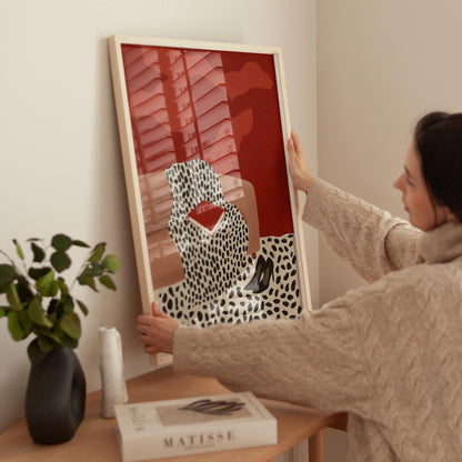 A woman is hanging a framed artwork on a wall, which features a red background with a black and white leopard print design.