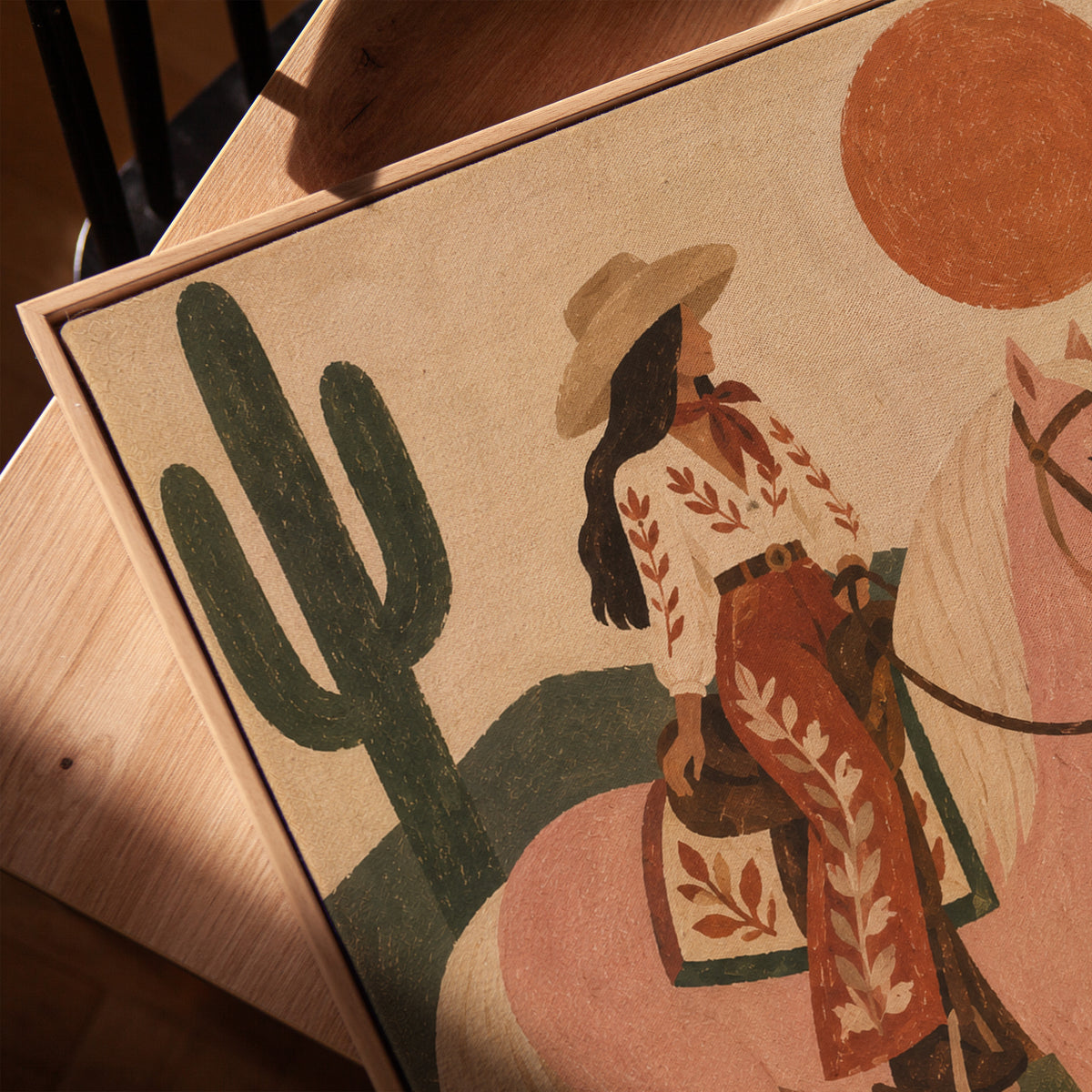 Cowgirl riding a pale rose horse past cacti under a red desert sun, vertical canvas print