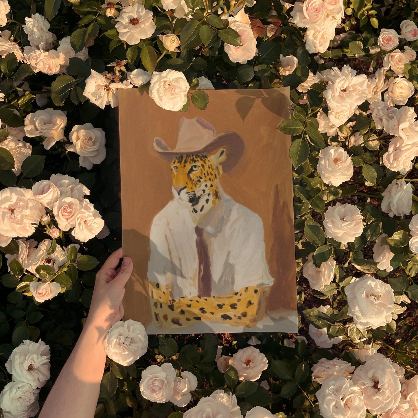 A hand holds a framed painting of a leopard wearing a cowboy hat, surrounded by a bed of white roses.