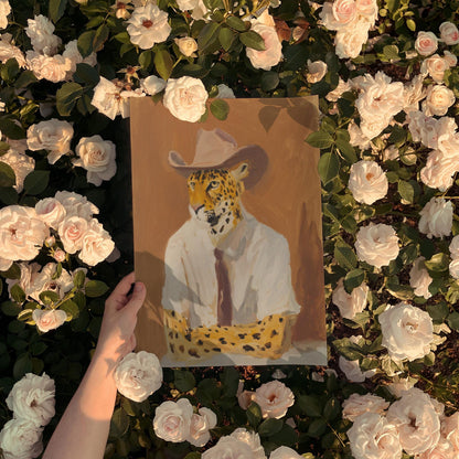 A hand holds a framed painting of a leopard wearing a cowboy hat, surrounded by a bed of white roses.
