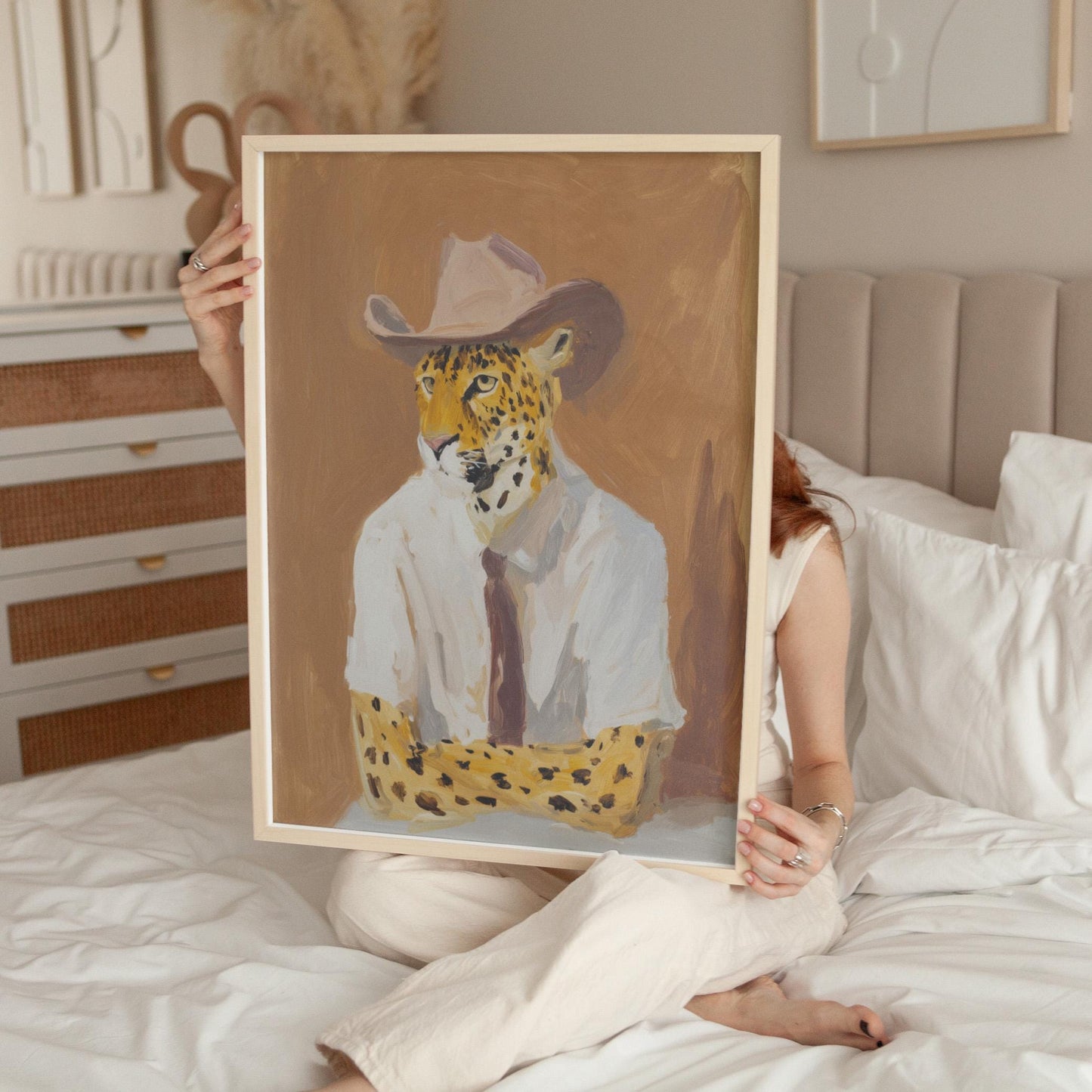 A person is sitting on a bed holding up a framed painting of a leopard wearing a white shirt and cowboy hat.