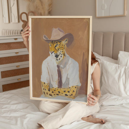 A person is sitting on a bed holding up a framed painting of a leopard wearing a white shirt and cowboy hat.