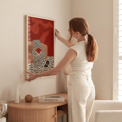 A woman in a white shirt and pants is hanging a framed artwork on a wall in a room with a wooden table and other decor items.