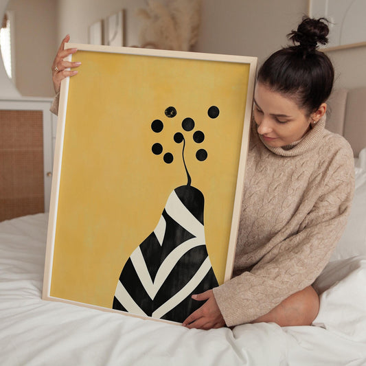 A woman is sitting on a bed and holding up a framed print with a yellow background and a black and white abstract design.