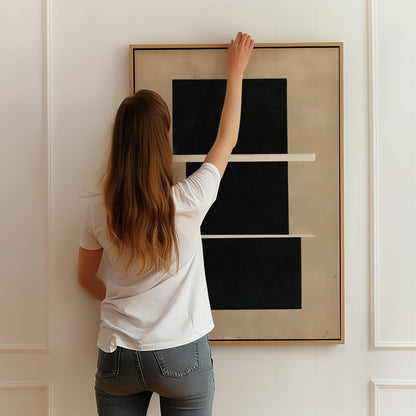 A woman with long brown hair is standing in front of a large black and white abstract painting, reaching up to hang it on a white wall.