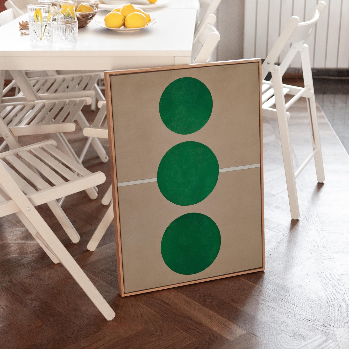 a framed artwork with a geometric design, including three green circles, placed on a wooden floor in a room with white chairs and a dining table.
