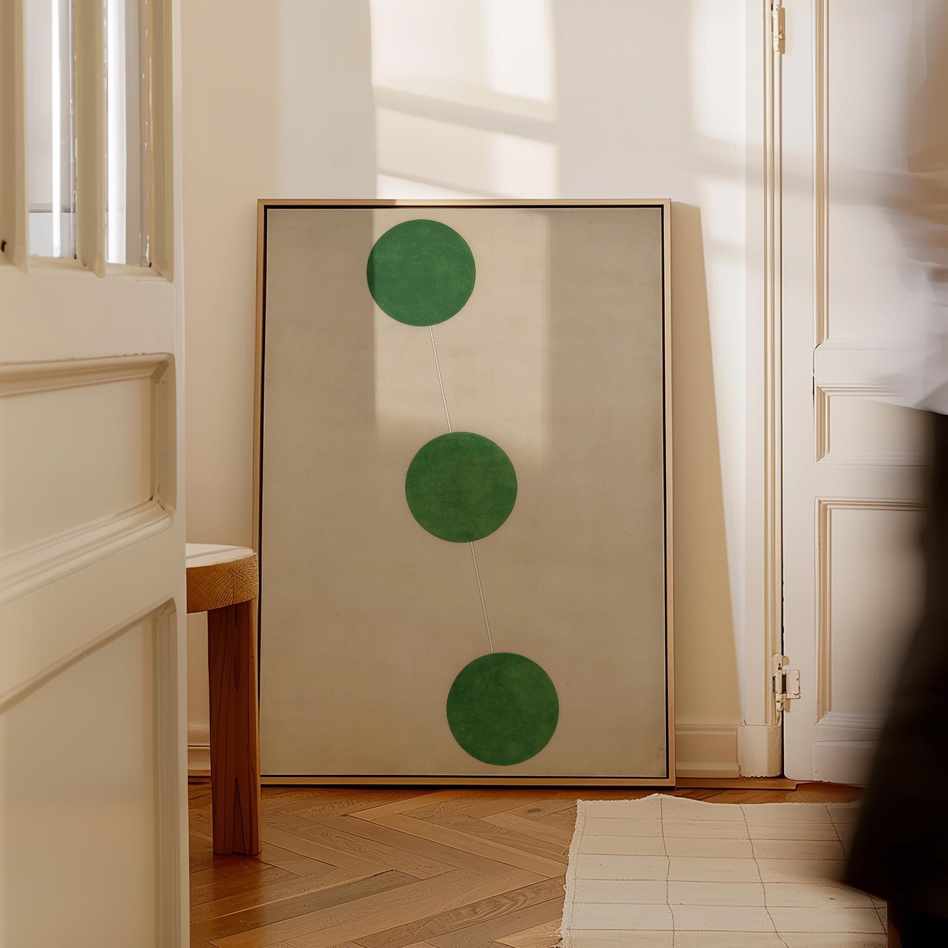 a large framed artwork with three green circles on a white background, placed in a hallway with a wooden floor.