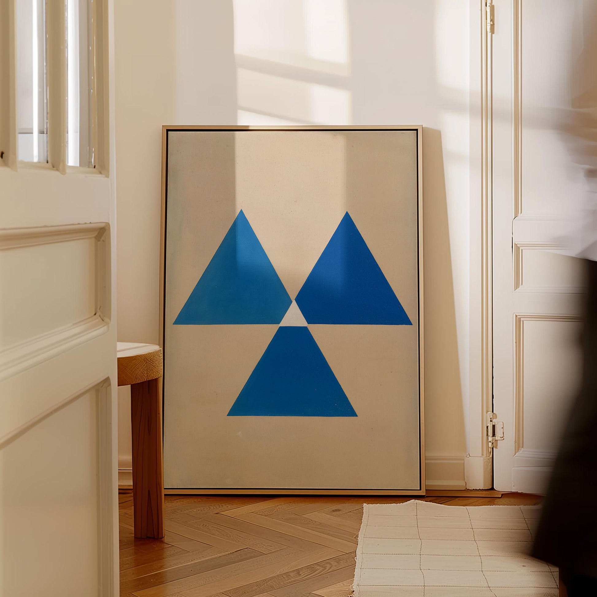a large framed artwork with a blue triangle design on a white background, placed in a room with a wooden floor and a white door.