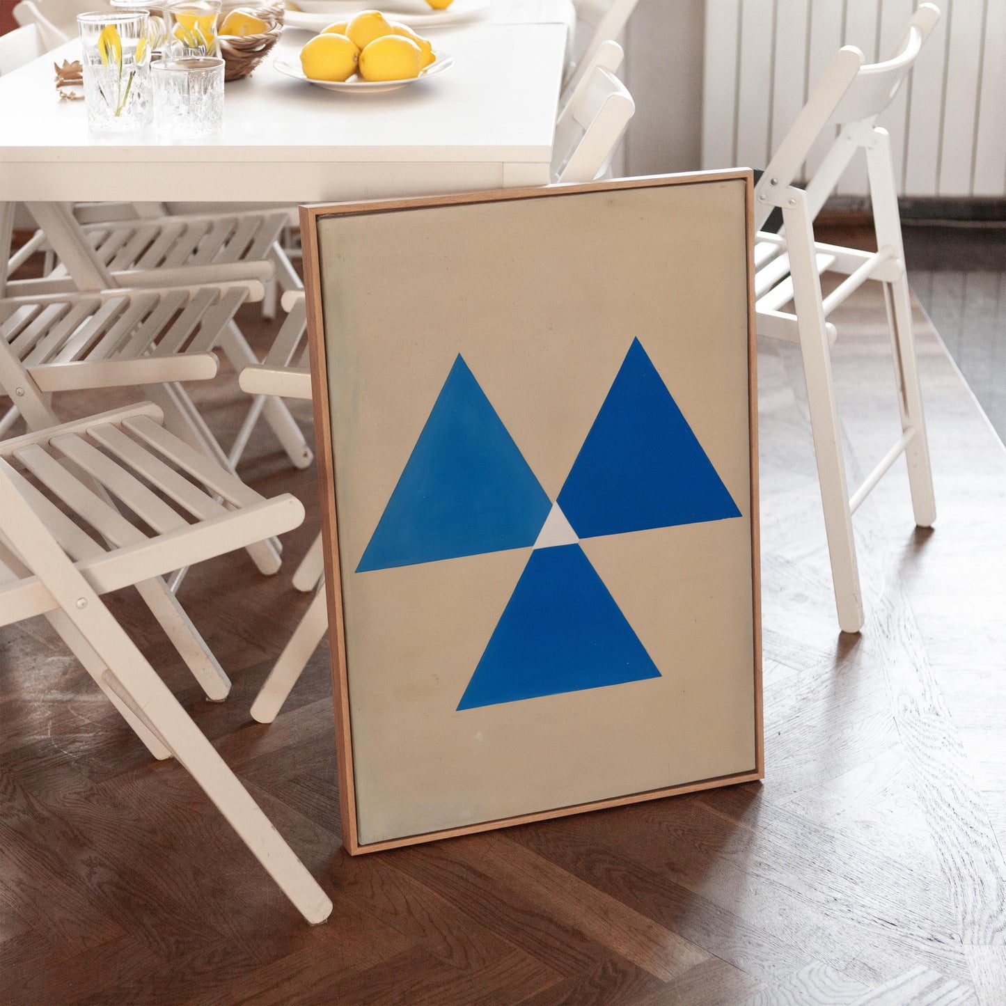 a framed artwork with a blue triangle design on a wooden floor, surrounded by white folding chairs and a dining table with a bowl of fruit.