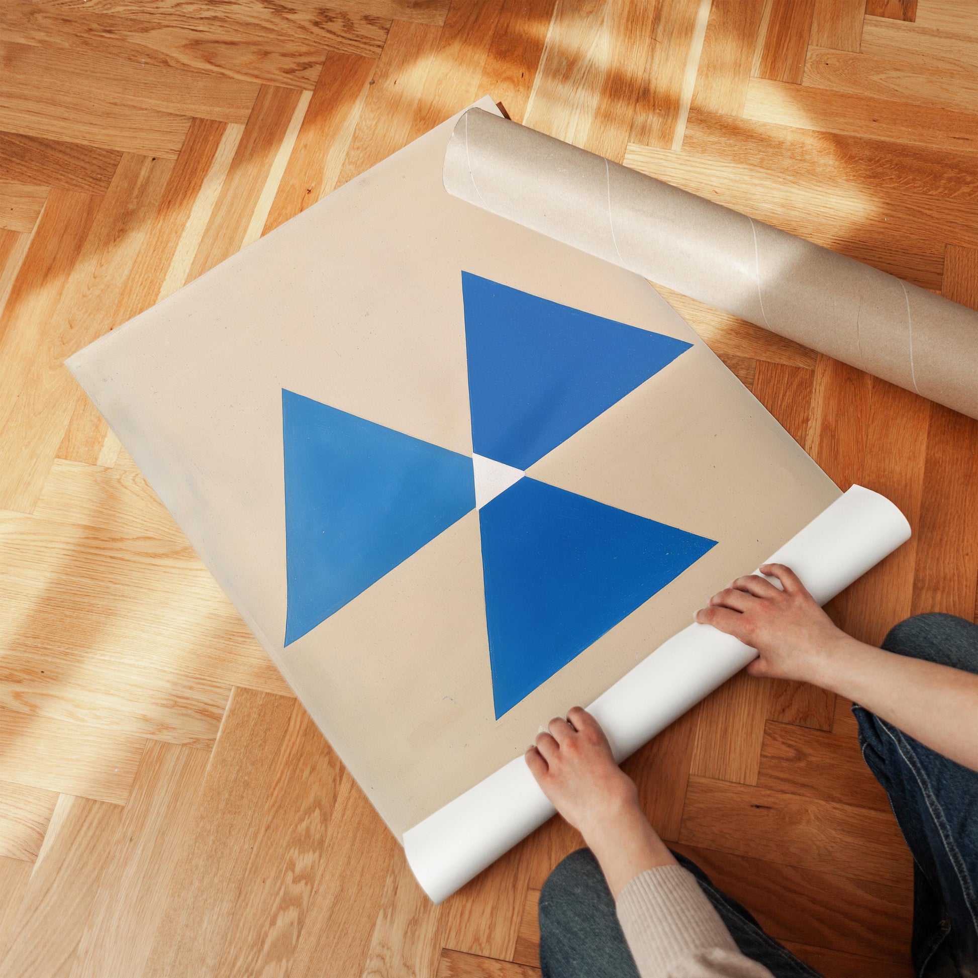 A person is rolling up a large piece of paper with a blue triangle design on it, which is placed on a wooden floor.