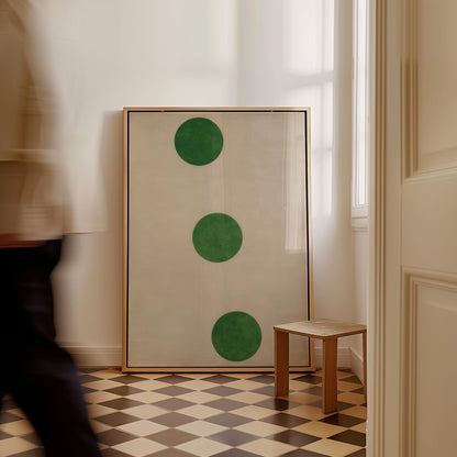 a large framed artwork with three green circles on a white background, placed in a room with a checkered floor and a small wooden stool.