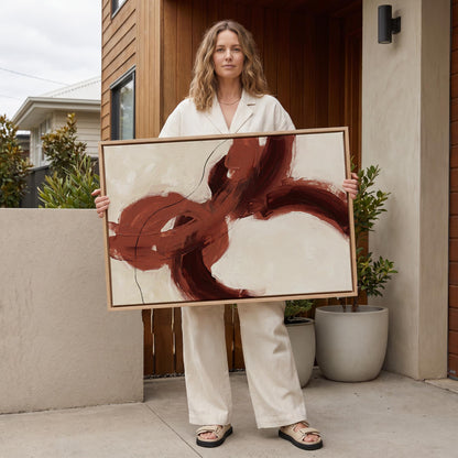A woman in a white outfit stands holding a large abstract painting with bold red and brown brushstrokes.
