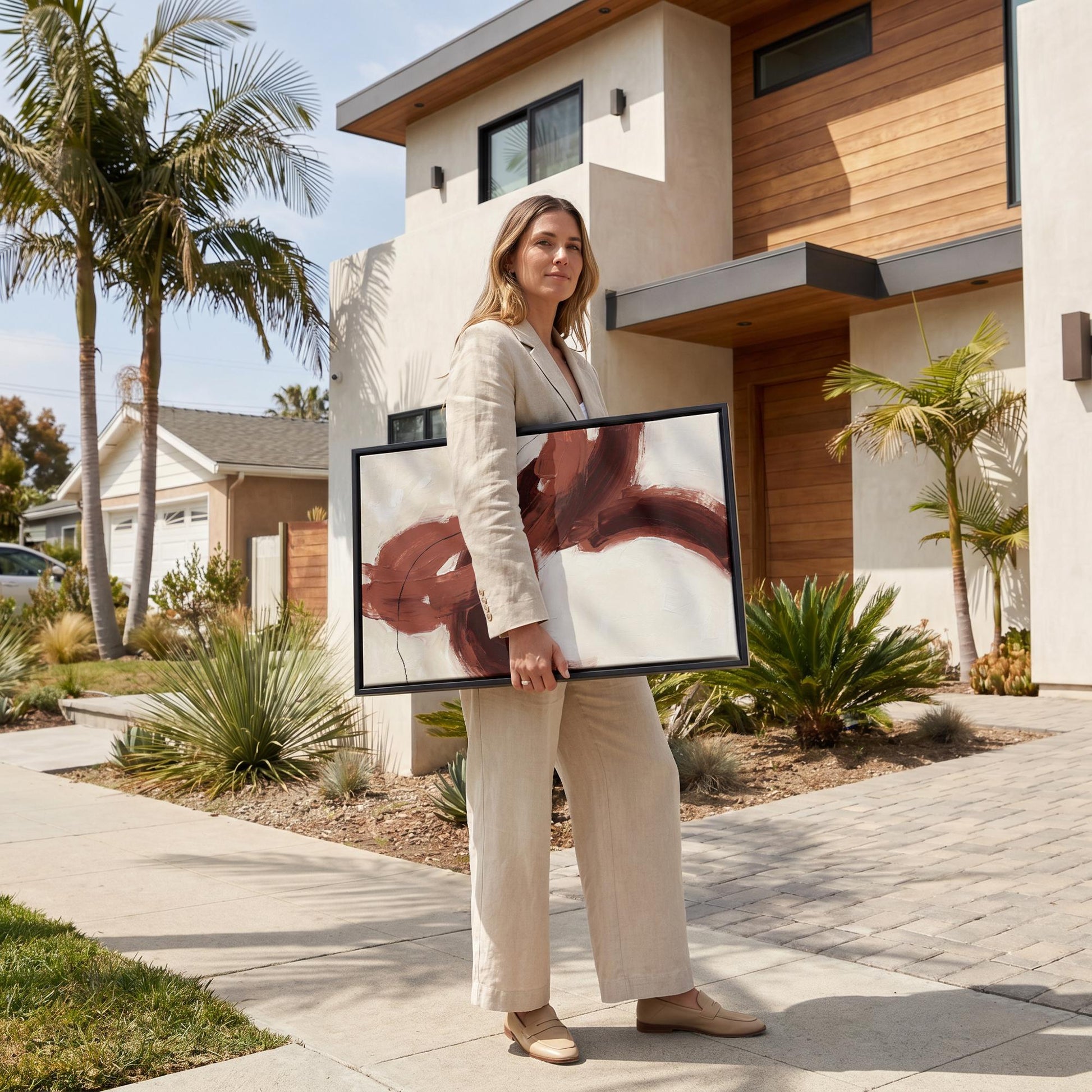 A woman in a beige suit is standing on a sidewalk, holding a large framed artwork in front of a modern house with a palm tree.