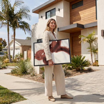 A woman in a beige suit is standing on a sidewalk, holding a large framed artwork in front of a modern house with a palm tree.