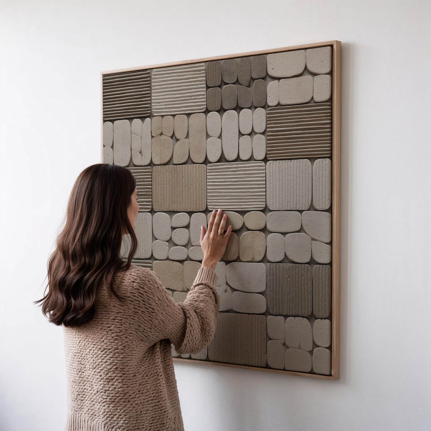 A woman in a brown sweater is standing in front of a large abstract wall art piece composed of various shapes and textures, including squares, rectangles, and circles.