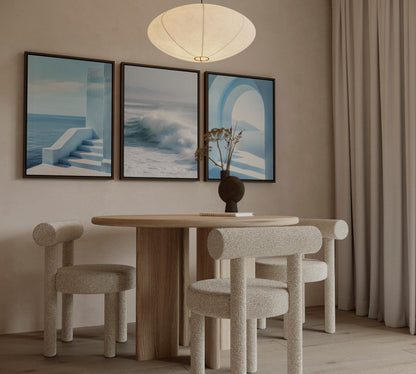 a modern dining room with a round wooden table and three beige chairs. Above the table, there are three framed pictures on the wall, depicting a staircase, a wave, and an archway. A pendant light hangs above the table, illuminating the space.