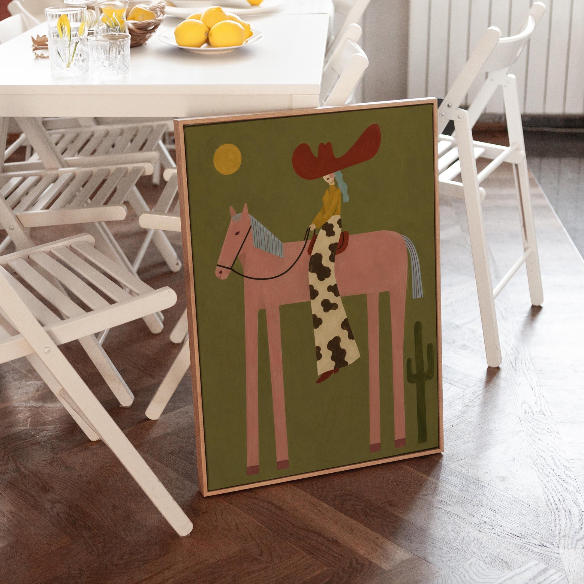 Cowgirl in a red hat riding a long-legged pink horse past a cactus at sunset, canvas wall art on a quiet wall in a considered interior