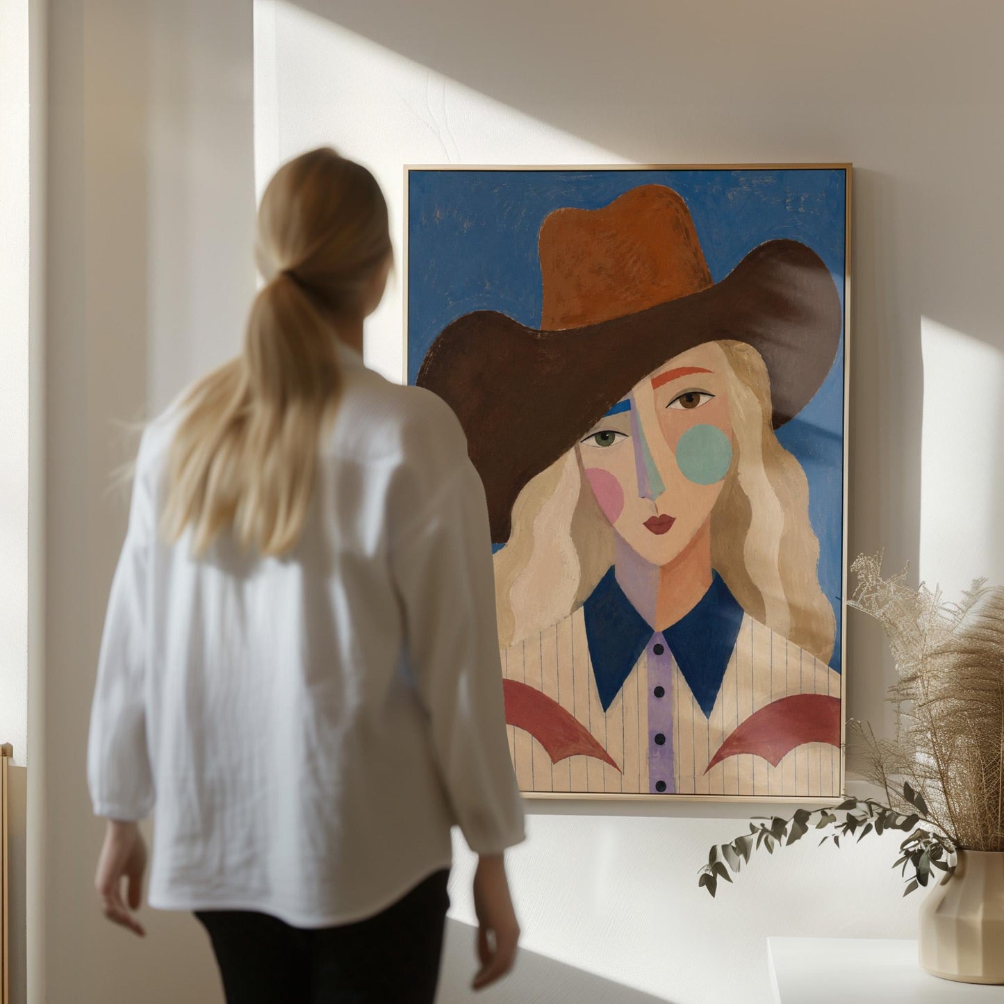 Blonde cowgirl in a rust hat with a cubist pastel face on deep blue, canvas print presented in a light, minimal room