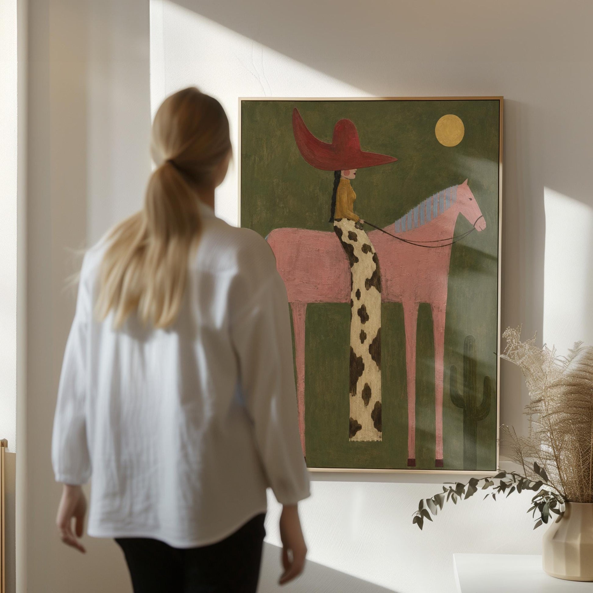 Cowgirl in a red hat riding a tall pink horse past a cactus under the moon, canvas print presented in a light, minimal room