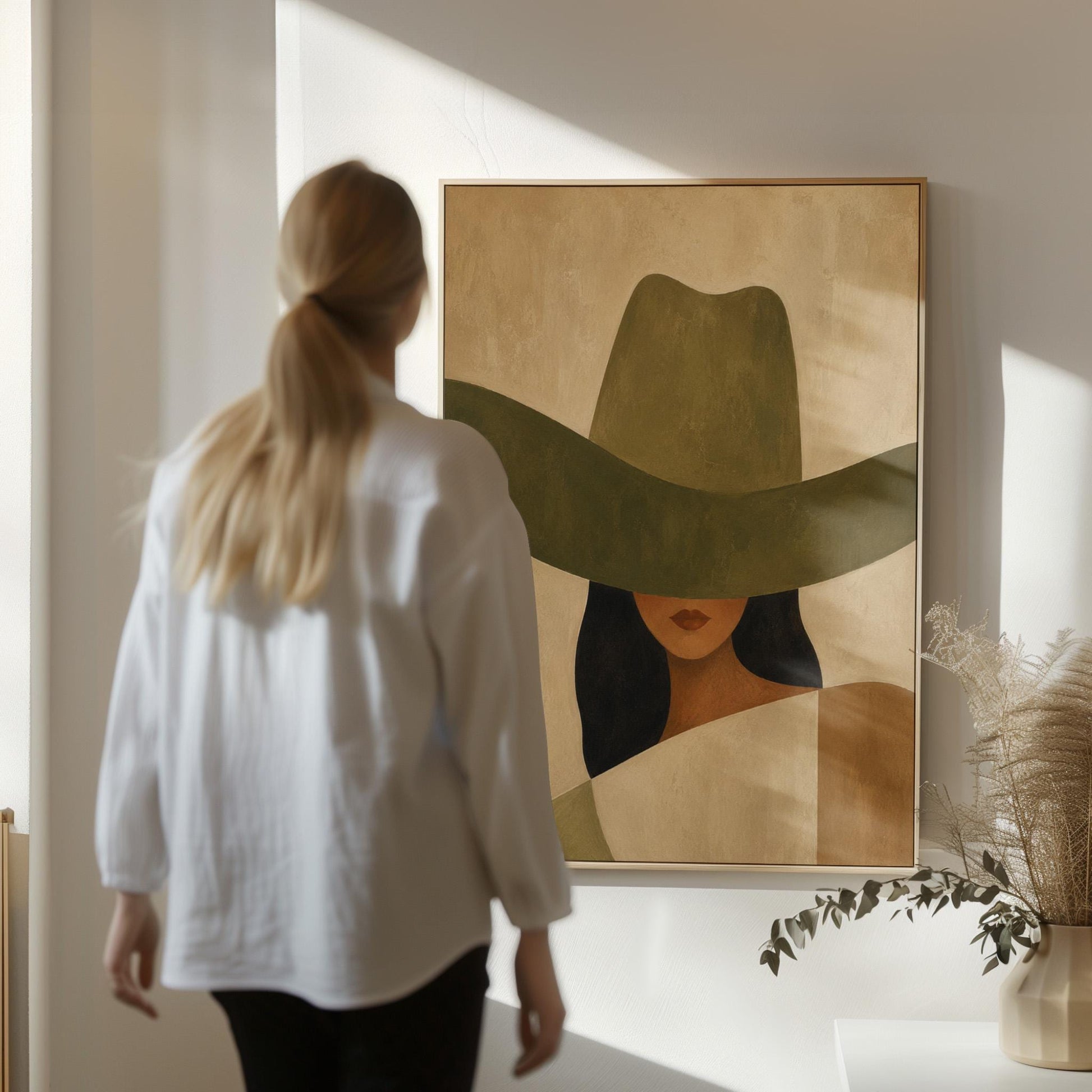 Cropped cowgirl face beneath the sweeping brim of a large olive hat, canvas print shown to scale in a furnished room
