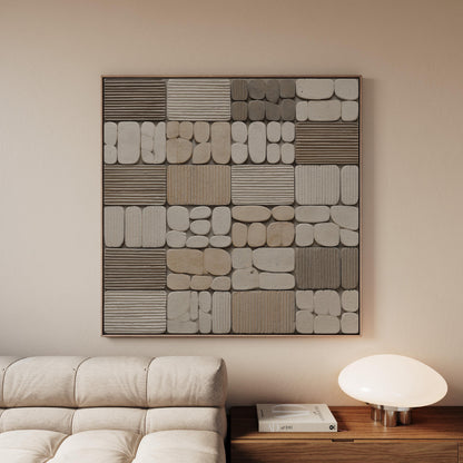 a modern living room with a large abstract artwork hanging on the wall above a white sofa. The artwork consists of various shades of gray and beige tiles arranged in a grid-like pattern. Next to the sofa, there is a wooden side table with a lamp on top, providing a warm and cozy atmosphere in the room.