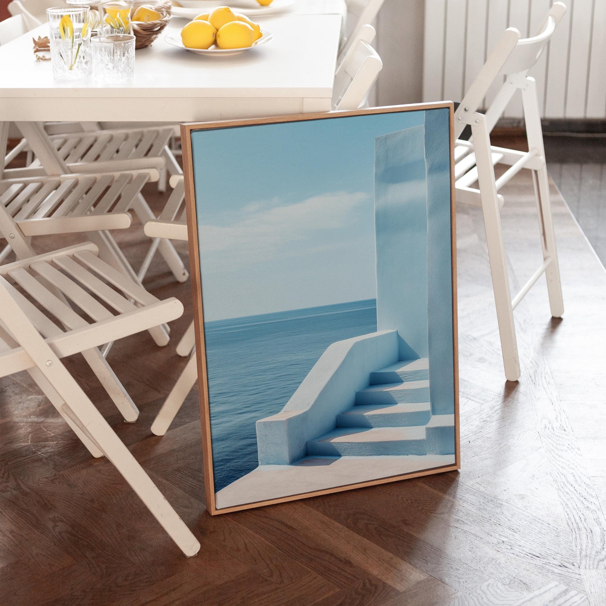 a framed photograph of a blue staircase leading to a serene ocean view, placed on a wooden floor in a room with white chairs and a table.