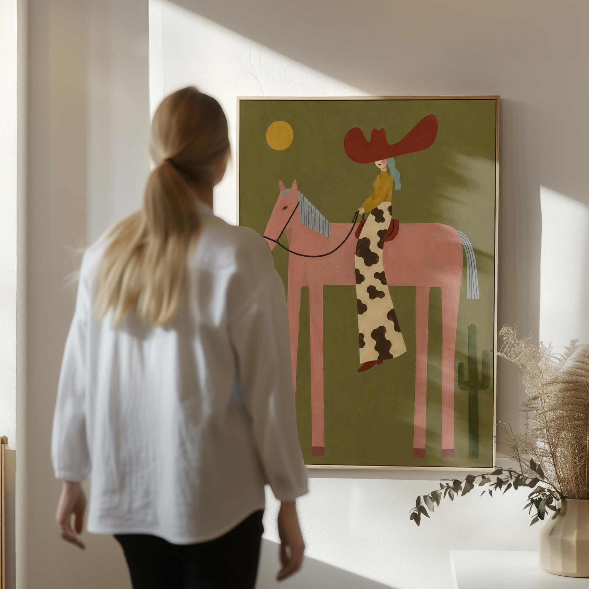 Cowgirl in a red hat riding a long-legged pink horse past a cactus at sunset, canvas print seen in a warm interior with natural light