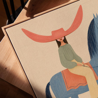 Cowgirl in a dramatic red hat riding a slender blue horse across pale sand, canvas print in a warm, minimally styled interior