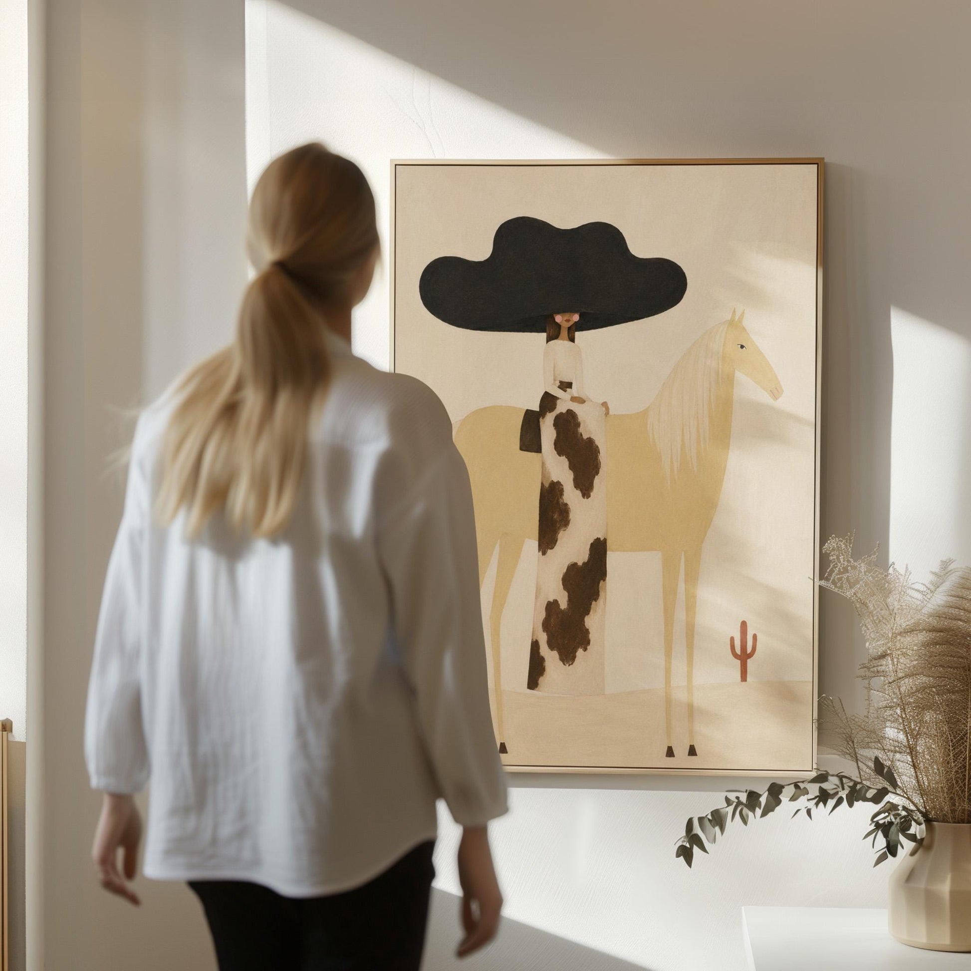 Woman in a huge black hat riding a pale palomino across a desert with cactus, canvas wall art seen in a warm interior with natural light