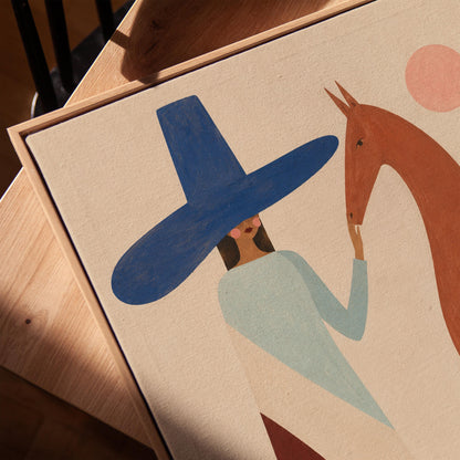 Woman in a tall blue hat reaching to a slim chestnut horse under a pink sun, canvas print in a warm, minimally styled interior