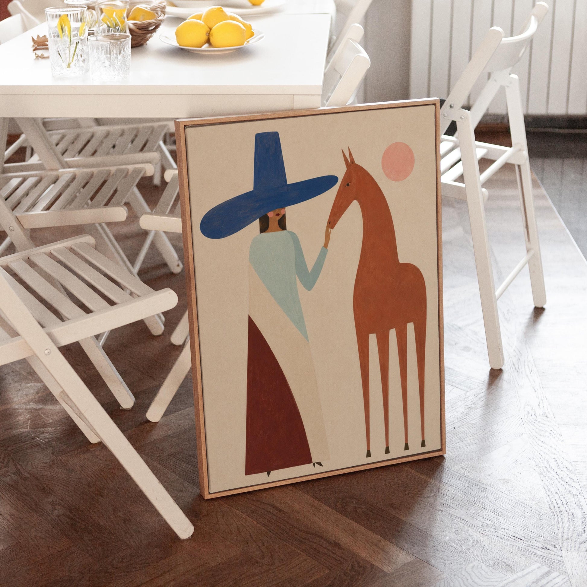 Woman in a tall blue hat reaching to a slim chestnut horse under a pink sun, canvas print styled against a soft white wall