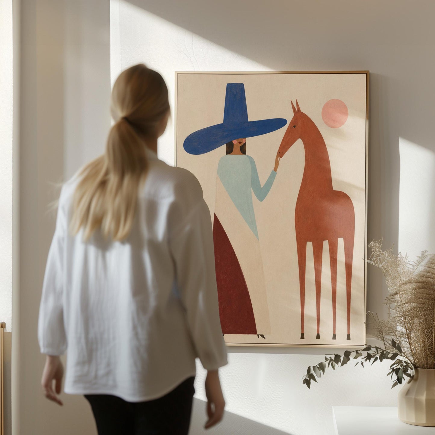 Woman in a tall blue hat reaching to a slim chestnut horse under a pink sun, canvas print hanging in a calm, modern living space