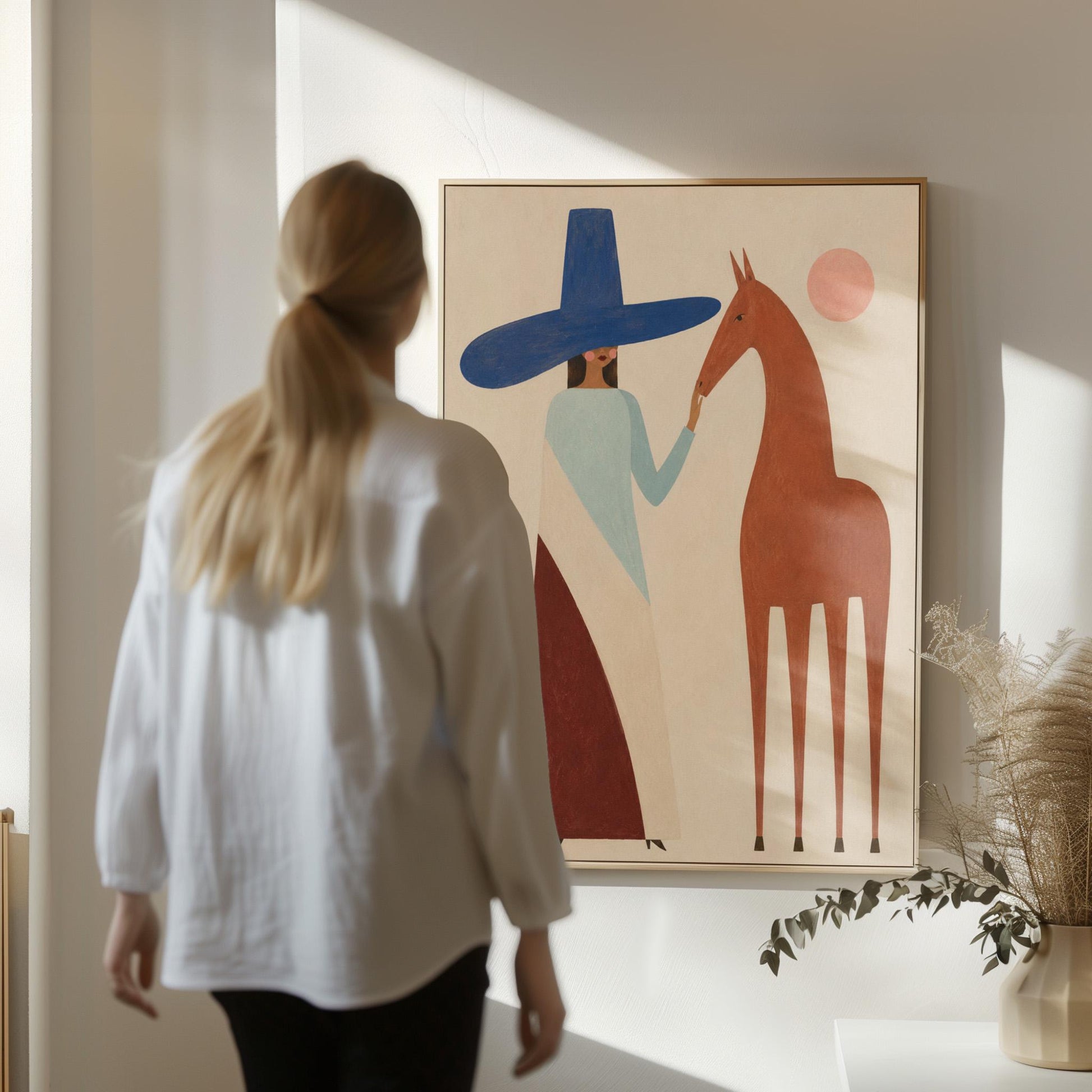 Woman in a tall blue hat reaching to a slim chestnut horse under a pink sun, canvas print hanging in a calm, modern living space