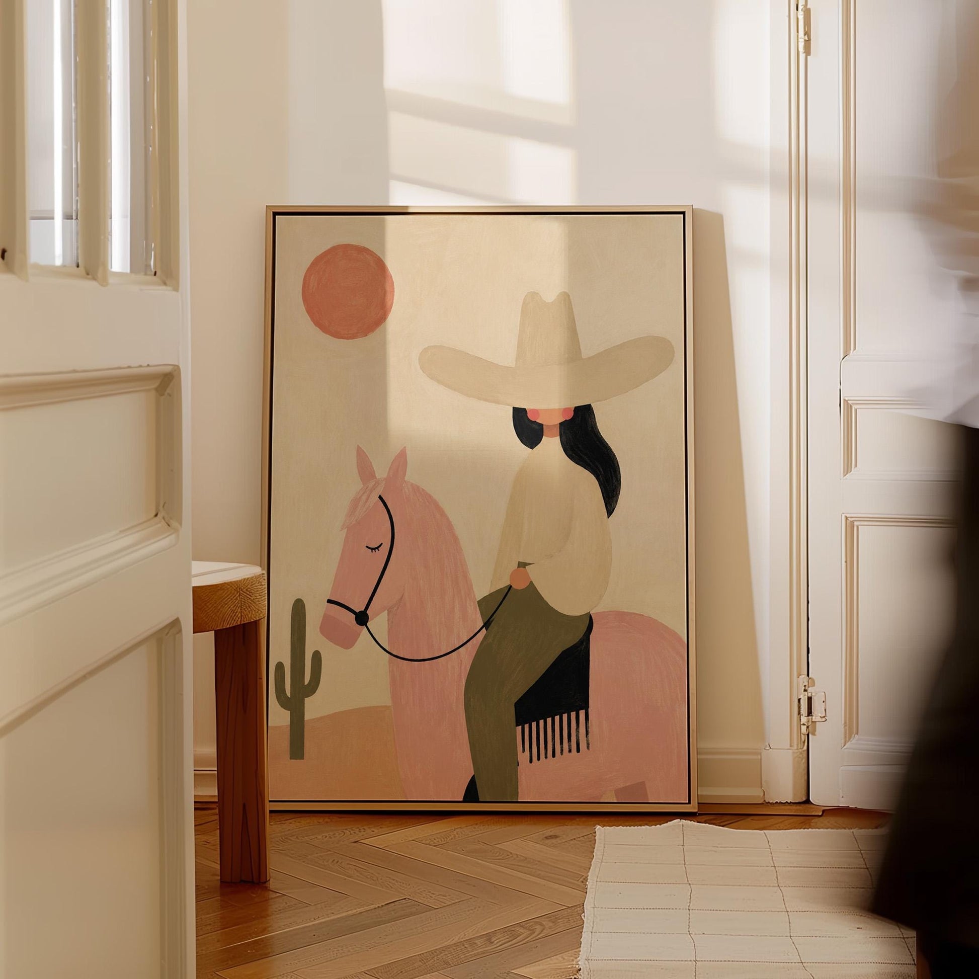 Cowgirl in a pale hat riding a soft pink horse past a desert cactus, canvas print shown to scale in a furnished room