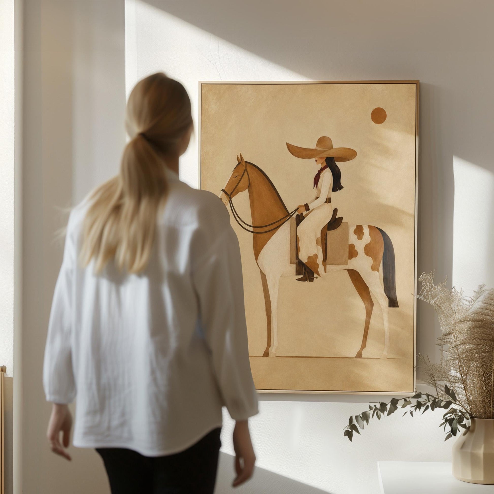 Cowgirl in a broad straw hat riding a cream and chestnut pinto horse, canvas print displayed on a neutral wall in a styled interior