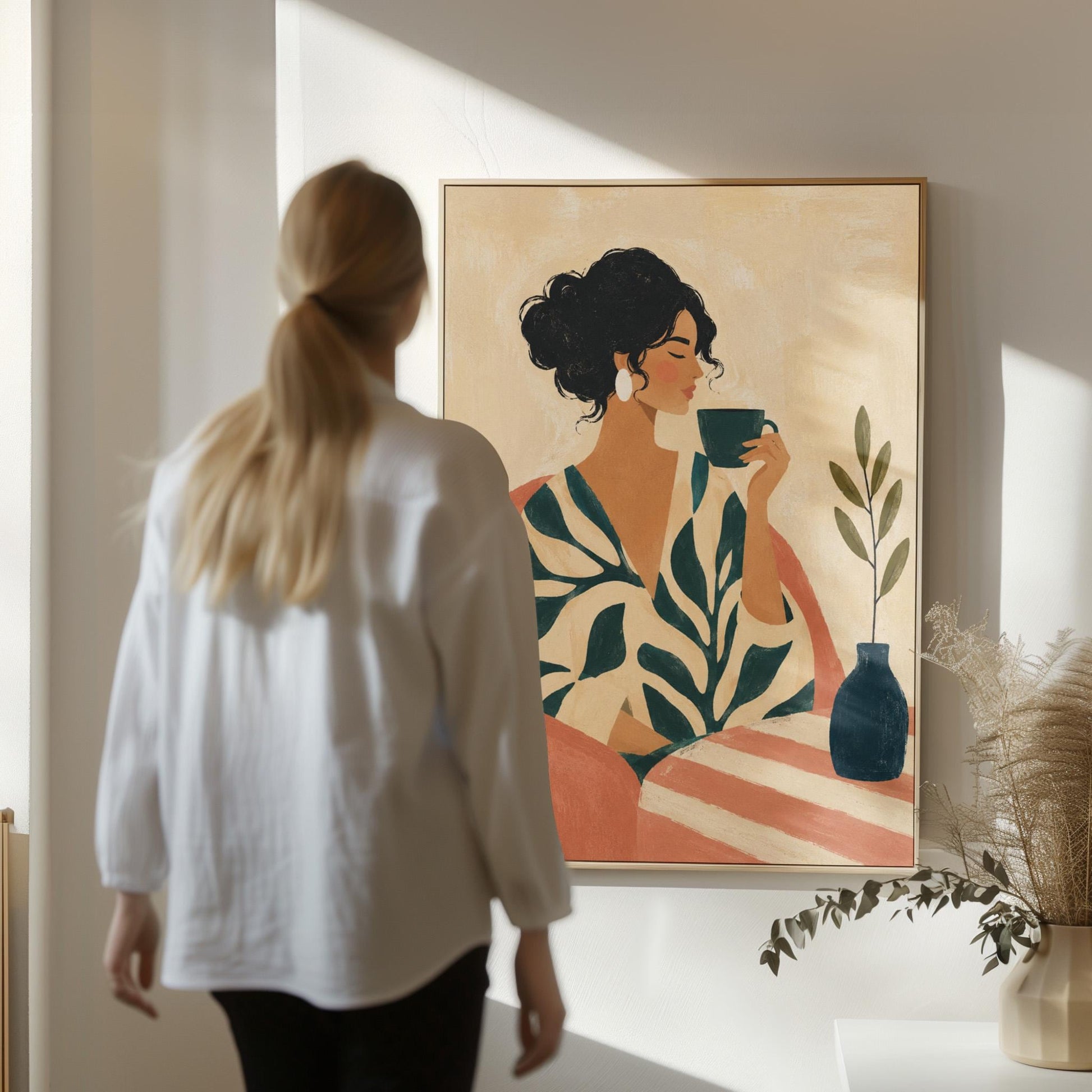 Woman in a leaf print robe drinking from a teal cup beside a vase, canvas wall art hanging in a calm, modern living space
