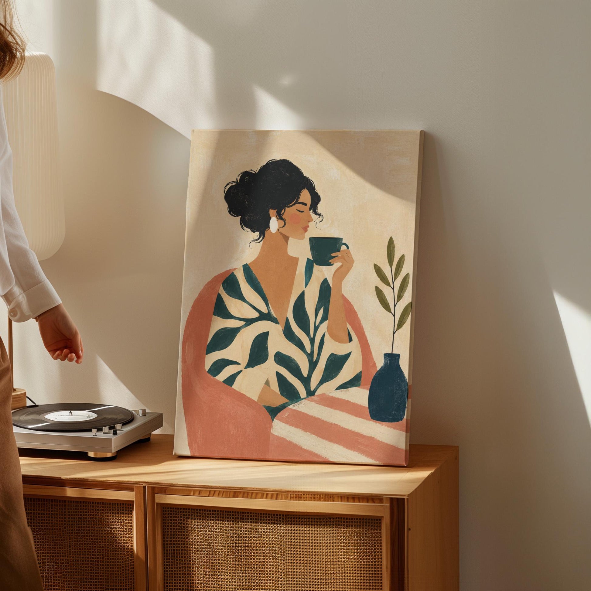 Woman in a leaf print robe drinking from a teal cup beside a vase, canvas wall art presented in a light, minimal room